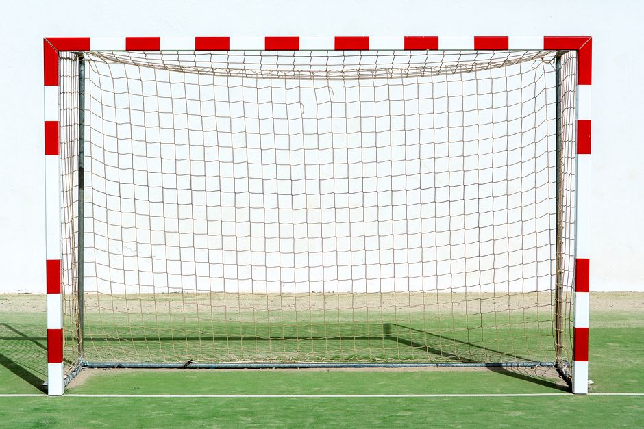 A solitary soccer goal with red and white colors on an outdoor sports field against a plain background.