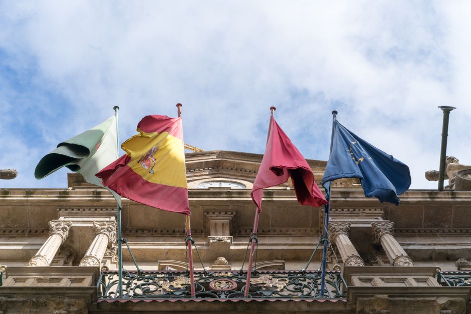 From below of Spanish flag and flags of Spanish provinces together with European Union flag waving on facade of magnificent stone building on sunny day