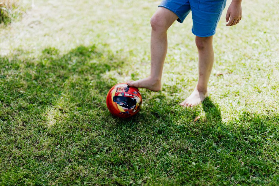 A young child in blue shorts plays barefoot with a red soccer ball on green grass.