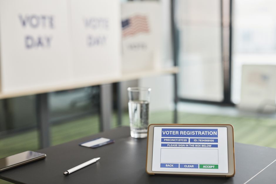Tablet displaying digital voter registration at indoor voting station with vote day signs.