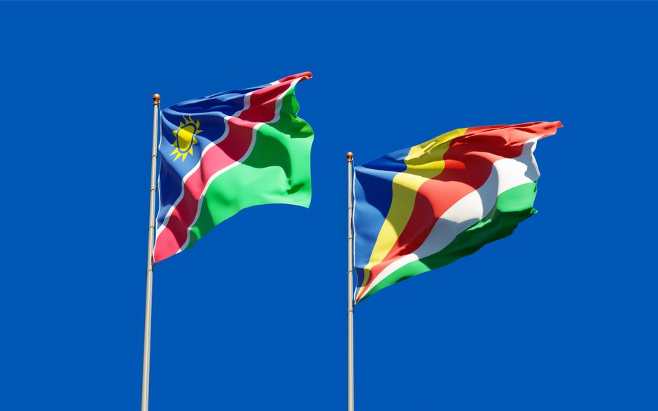 Colorful flags of Namibia and Seychelles waving on flagpoles against a clear blue sky.